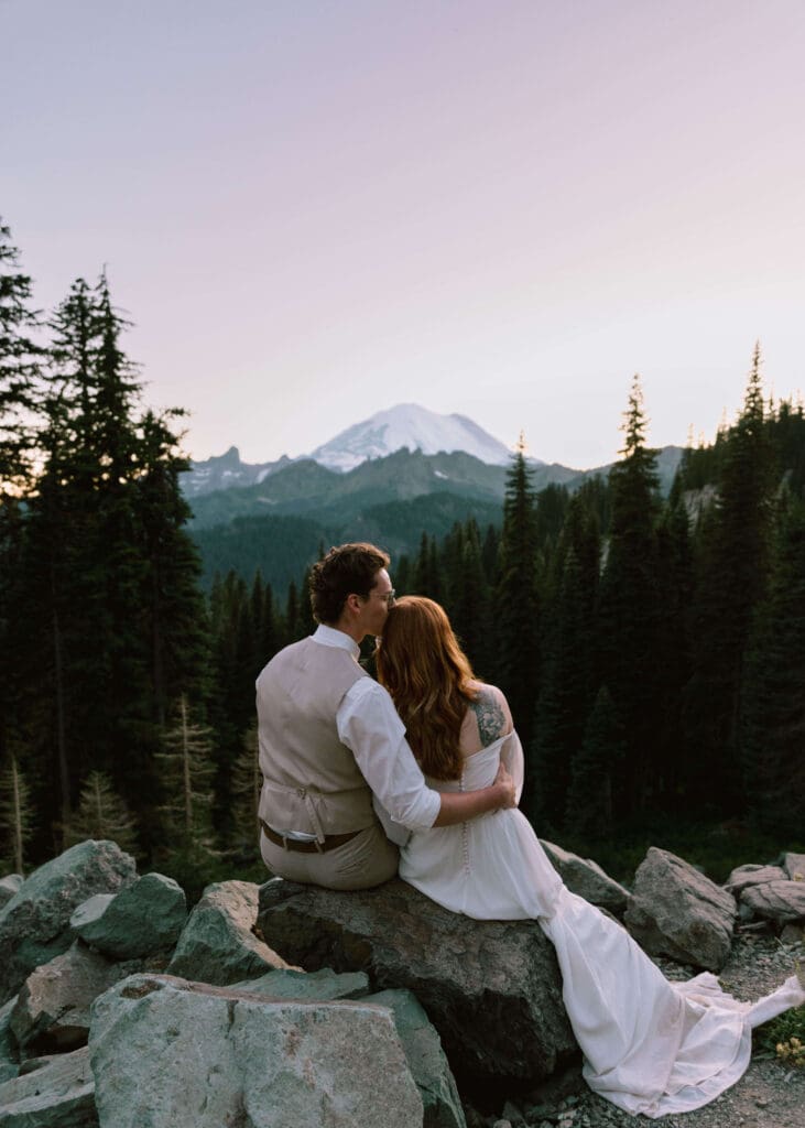 groom kissing top of his bride's head while sitting on a rock looking at Mt Rainier during blue hour 