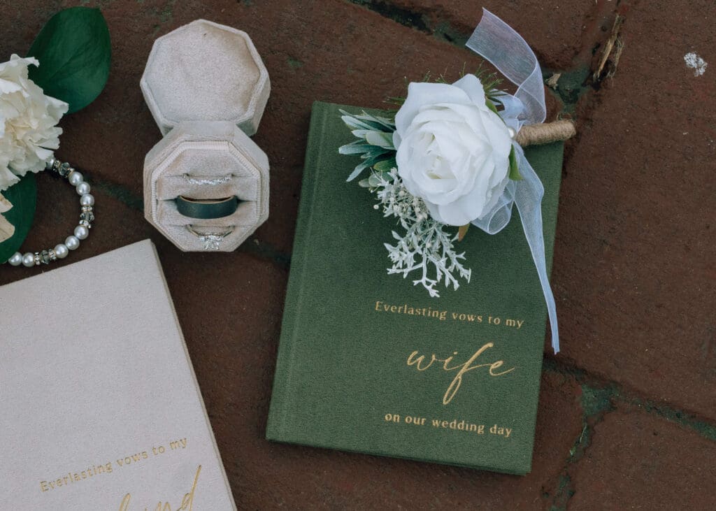 green vow book with white rose boutonniere and rings in ring box on the brick path at Pickering Barn