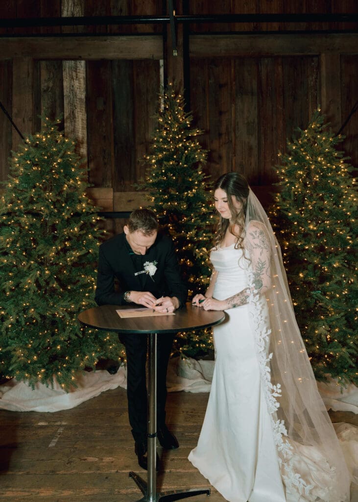 bride and groom signing their marriage license inside wedding venue in front of lit christmas trees