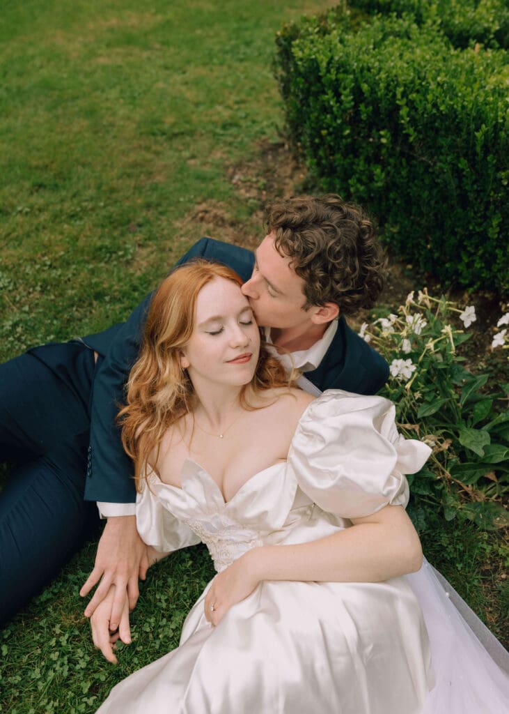 bride and groom laying in the grass with groom kissing the side of her head