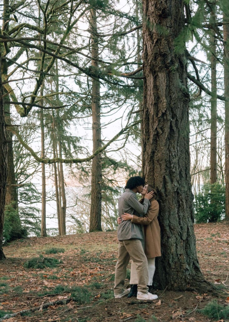 man and woman kissign against a tree in the forest with an ocean view at susnet at Point Defiance