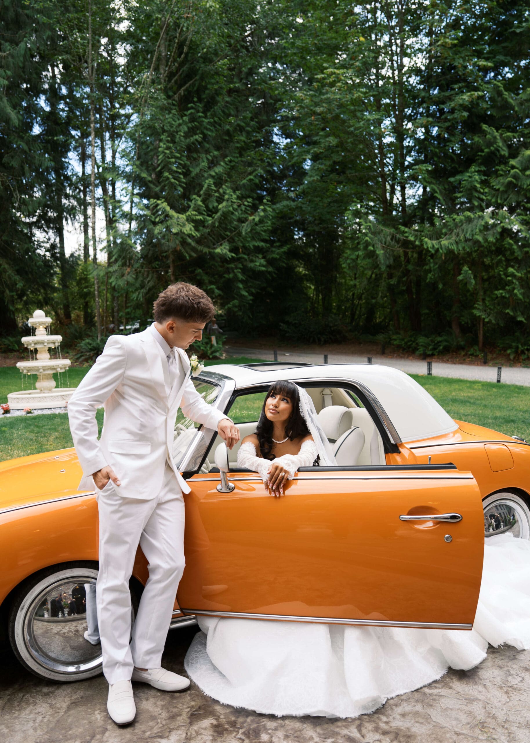 groom in white leaning against orange vintage car looking at bride sitting in the car at Washington wedding