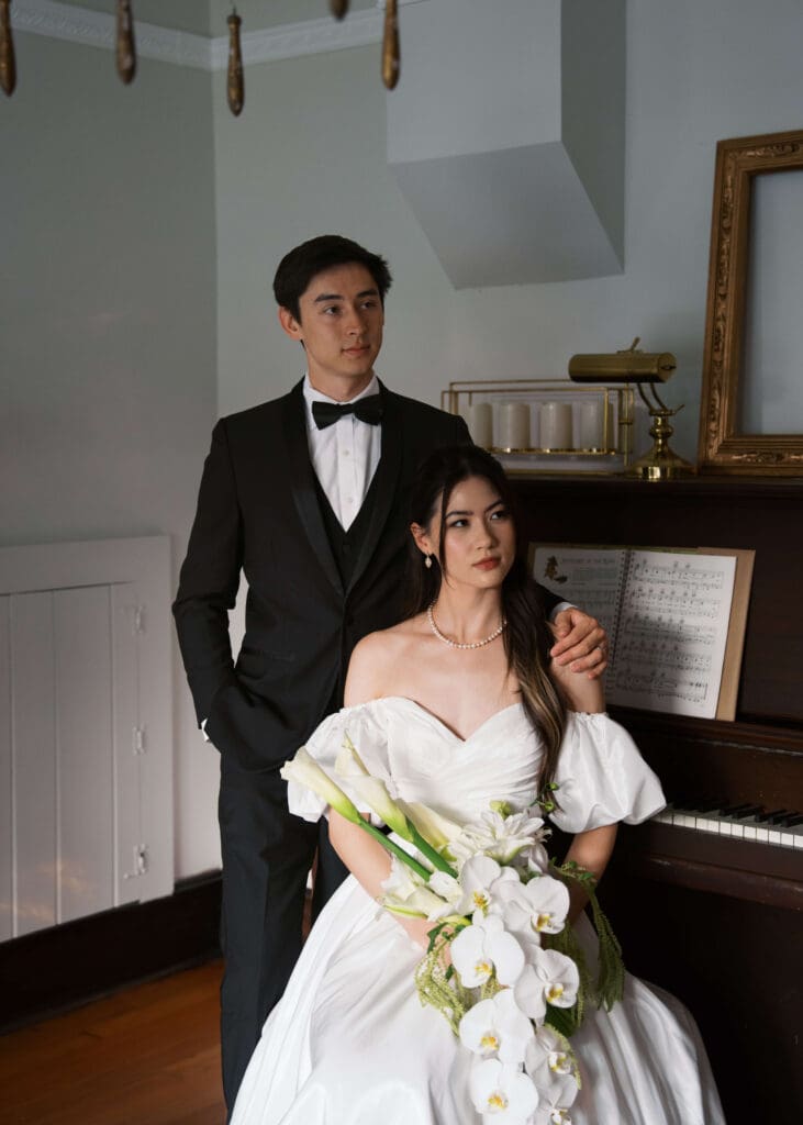 editorial posed couple with bride sitting on piano bench with her bouquet and groom standing behind her at