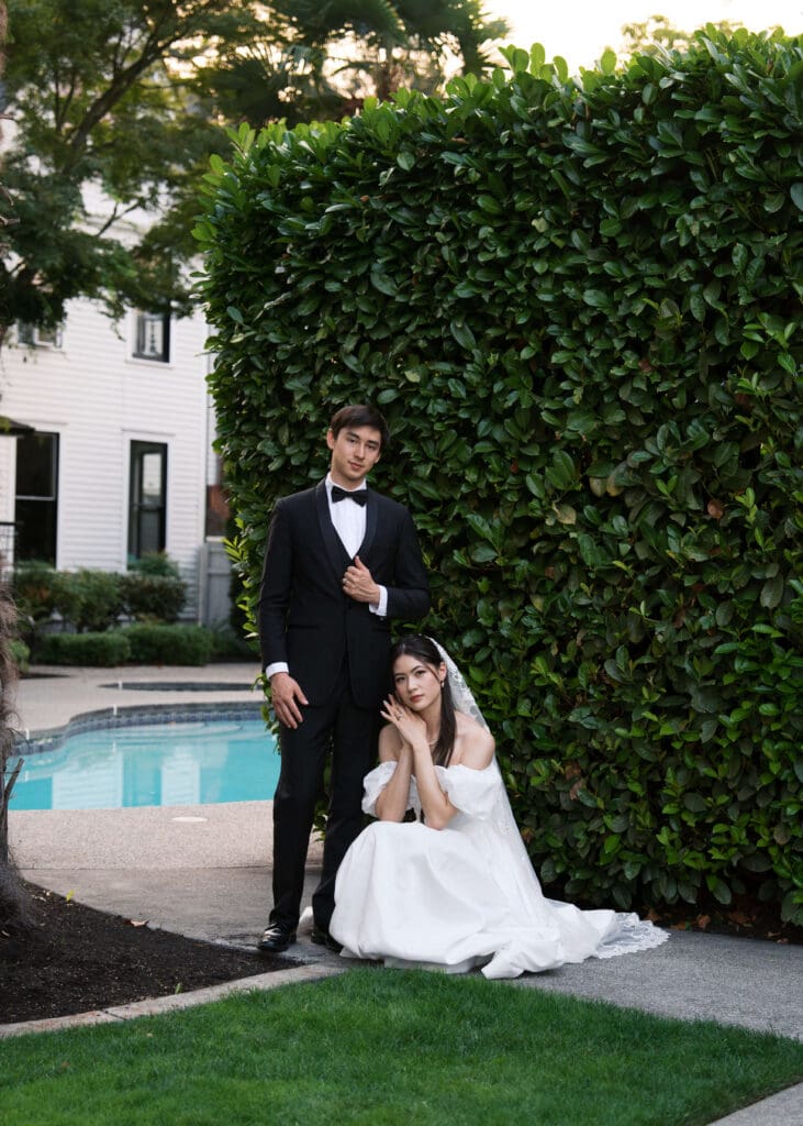 groom standing and holding his suit coat while his bride squats in front of him in an editorial pose in front of the pool at Orting Manor