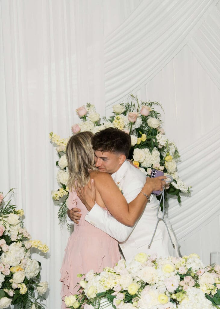 groom in white hugging his mom against a white backdrop at wedding