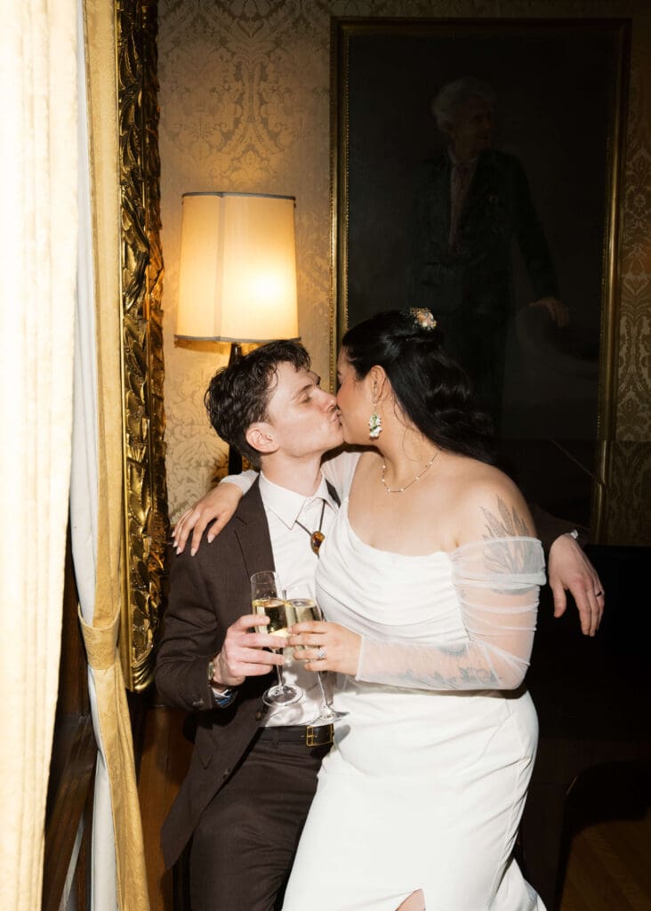 bride and groom kissing while tapping champagne glasses and sitting on a piano bench at Stimson-Green Mansion