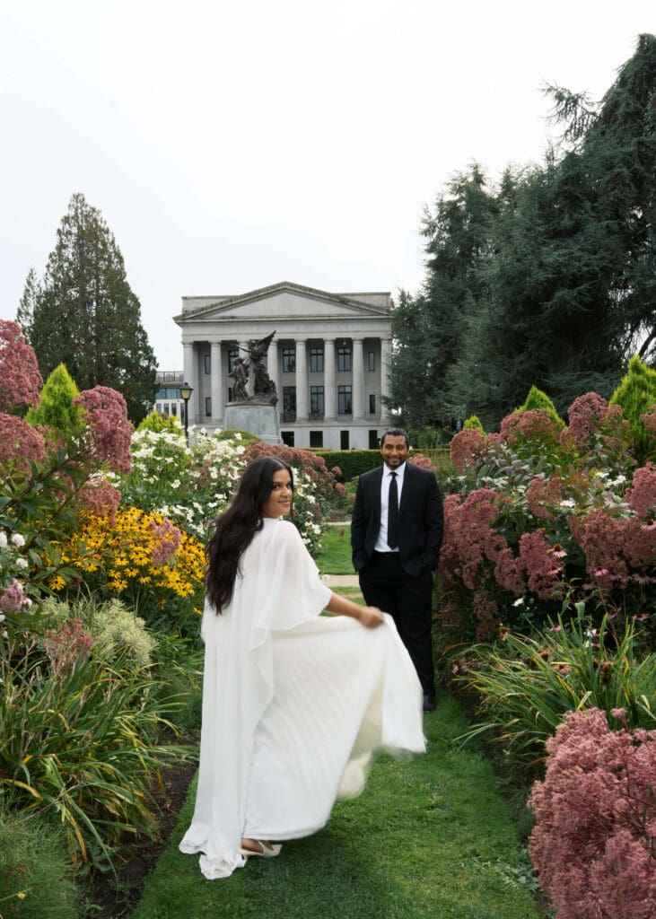 bride holding dress and looking over her shoulder while groom admires her in the garden at the Washington State Capitol wedding